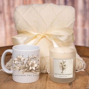 May include: A cream-colored blanket with a satin ribbon bow, a white mug with a floral design and the text "Consider the Lilies", and a candle with a floral design and the text "Seek First His Kingdom".