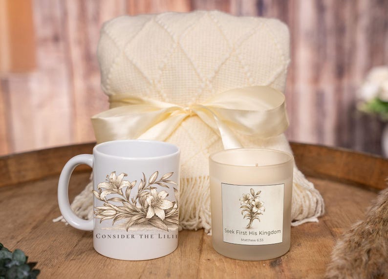 May include: A cream-colored blanket with a satin ribbon bow, a white mug with a floral design and the text "Consider the Lilies", and a candle with a floral design and the text "Seek First His Kingdom".