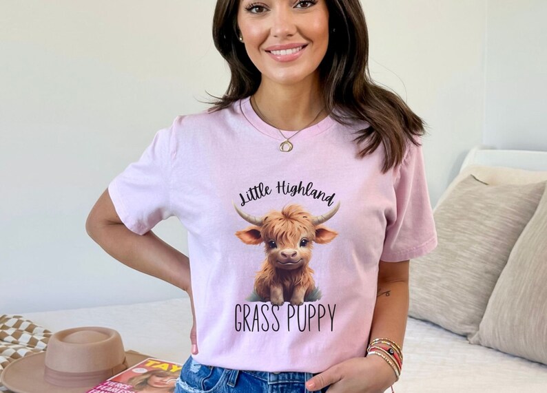 May include: A pink t-shirt featuring a cartoon Highland cow with the text "Little Highland Grass Puppy".