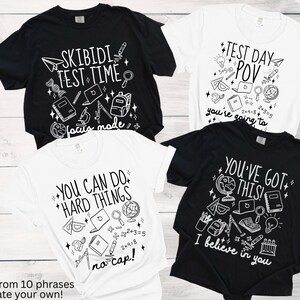 Puede incluir: Cuatro camisetas, dos negras y dos blancas, con gr&aacute;ficos y texto con tem&aacute;tica escolar. Las camisetas presentan frases como "Skibidi Test Time" y "You've Got This!" con ilustraciones de &uacute;tiles escolares. El texto "choose from 10 phrases or create your own!" est&aacute; en la parte inferior.