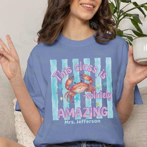 May include: A periwinkle blue t-shirt featuring a graphic of a smiling orange crab centered over light blue vertical stripes. The text reads This class is -solutely AMAZING and Mrs. Jefferson in purple and white lettering with small stars.