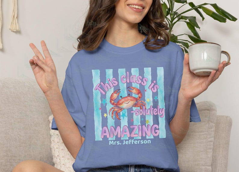 May include: A periwinkle blue t-shirt featuring a graphic of a smiling orange crab centered over light blue vertical stripes. The text reads This class is -solutely AMAZING and Mrs. Jefferson in purple and white lettering with small stars.
