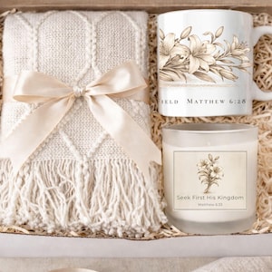 May include: A gift box containing a cream-colored throw blanket tied with a satin bow, a white mug with floral design and text, and a candle with a floral design and text. The mug and candle have the same floral design.