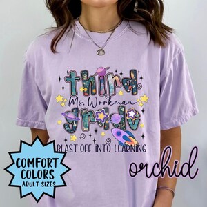 May include: A light purple short-sleeve t-shirt featuring a space-themed graphic with the text "third grade", "Ms. Workman", and "BLAST OFF INTO LEARNING". The design includes planets, stars, and a rocket ship in shades of blue, purple, and yellow.