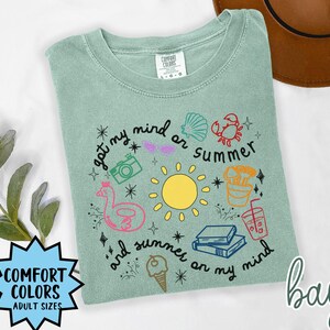 May include: A light green t-shirt with a graphic design featuring a sun, a beach umbrella, a camera, a seashell, a crab, a drink, a life preserver, and the text "got my mind on summer and summer on my mind".