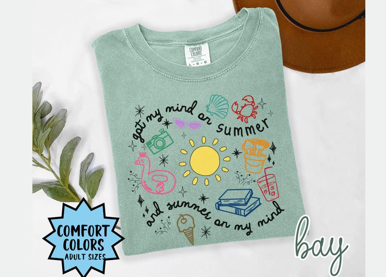 May include: A light green t-shirt with a graphic design featuring a sun, a beach umbrella, a camera, a seashell, a crab, a drink, a life preserver, and the text "got my mind on summer and summer on my mind".