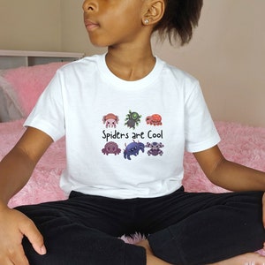Pode incluir: T-shirt branca com um gr&aacute;fico colorido de aranhas de desenho animado e o texto "Spiders are Cool". As aranhas s&atilde;o de v&aacute;rias cores e designs. A camisa &eacute; feita de um material macio.