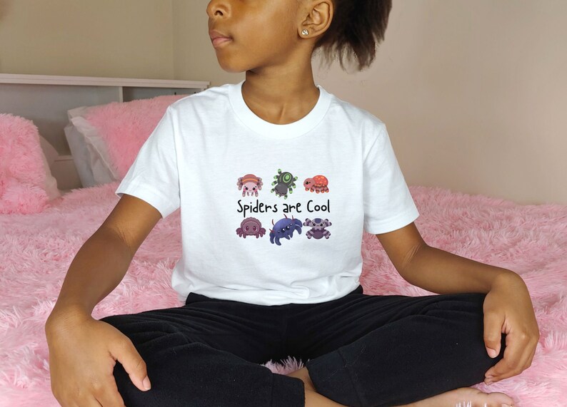 Pode incluir: T-shirt branca com um gr&aacute;fico colorido de aranhas de desenho animado e o texto "Spiders are Cool". As aranhas s&atilde;o de v&aacute;rias cores e designs. A camisa &eacute; feita de um material macio.