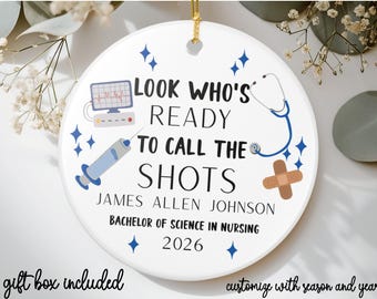 May include: A white ceramic ornament with a gold string for hanging. The ornament features medical-themed illustrations and text that reads "LOOK WHO'S READY TO CALL THE SHOTS." It includes a name, degree, and the year 2026.