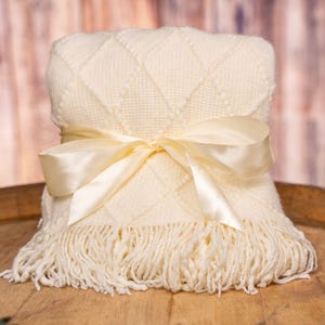 May include: A folded cream-colored blanket with a diamond pattern and fringe, tied with a satin ribbon. The blanket is displayed on a wooden surface, suggesting a cozy and inviting theme.