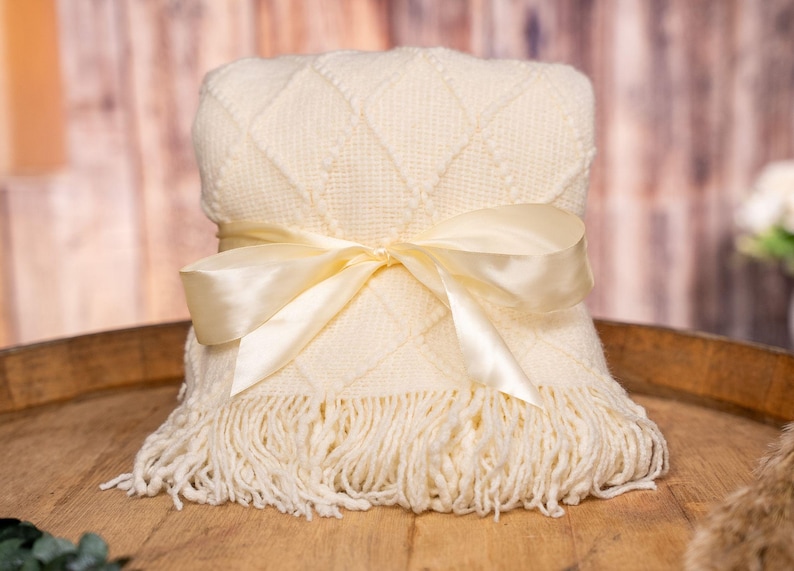 May include: A folded cream-colored blanket with a diamond pattern and fringe, tied with a satin ribbon. The blanket is displayed on a wooden surface, suggesting a cozy and inviting theme.