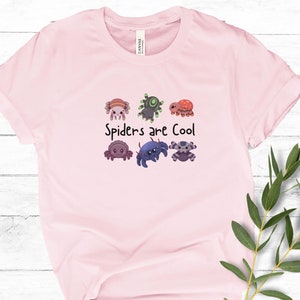 Pode incluir: T-shirt rosa claro com a frase "Spiders are Cool" e um desenho de aranhas de desenho animado. As aranhas t&ecirc;m cores e padr&otilde;es diferentes. A t-shirt &eacute; feita de um material macio.