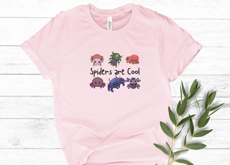 Pode incluir: T-shirt rosa claro com a frase "Spiders are Cool" e um desenho de aranhas de desenho animado. As aranhas t&ecirc;m cores e padr&otilde;es diferentes. A t-shirt &eacute; feita de um material macio.