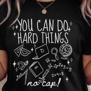 Puede incluir: Camiseta negra con la frase "YOU CAN DO HARD THINGS" en blanco y varios gr&aacute;ficos relacionados con la escuela, incluyendo un transportador, un port&aacute;til, un globo terr&aacute;queo y s&iacute;mbolos matem&aacute;ticos. Tambi&eacute;n incluye la frase "no cap!".