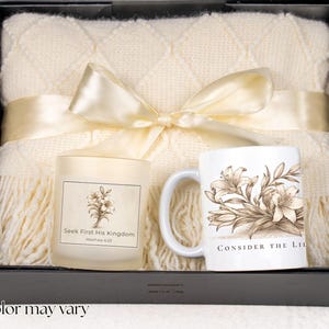 May include: A gift set in a black box, featuring a cream-colored throw blanket, a candle with floral design and text, and a white mug with a floral illustration and text. The blanket is tied with a cream ribbon.