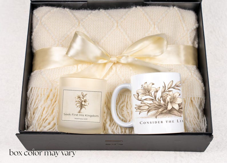 May include: A gift set in a black box, featuring a cream-colored throw blanket, a candle with floral design and text, and a white mug with a floral illustration and text. The blanket is tied with a cream ribbon.