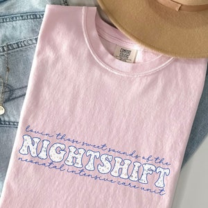 May include: A light pink Comfort Colors t-shirt with the text "Lovin' those sweet sounds of the NIGHTSHIFT neonatal intensive care unit" in a blue and white design. A tan hat and blue jeans are also in the image.