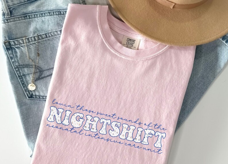 May include: A light pink Comfort Colors t-shirt with the text "Lovin' those sweet sounds of the NIGHTSHIFT neonatal intensive care unit" in a blue and white design. A tan hat and blue jeans are also in the image.