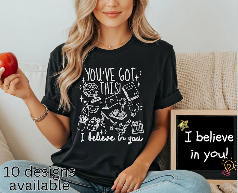 Puede incluir: Camiseta negra con el texto "YOU'VE GOT THIS! I believe in you" e ilustraciones escolares en blanco. Una peque&ntilde;a pizarra muestra "I believe in you!" con una estrella y una bombilla. La frase "10 designs available" est&aacute; en la parte inferior.