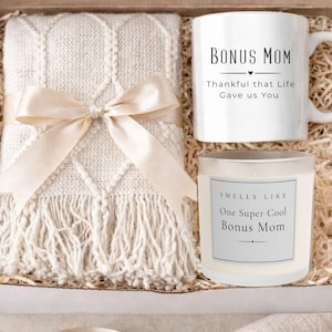 May include: A gift box containing a cream-colored blanket tied with a satin bow, a white mug with "BONUS MOM" text, and a candle labeled "Smells Like One Super Cool Bonus Mom". The box is filled with shredded paper.