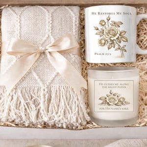 May include: A gift box containing a cream-colored throw blanket tied with a satin ribbon, a white mug with a floral design and the text "He Restores My Soul" and a candle with the text "He Guides Me Along the Right Paths".