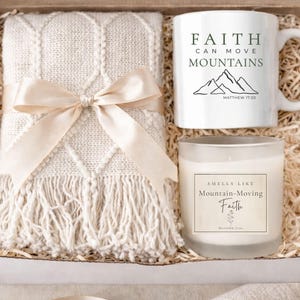 May include: A gift box containing a cream-colored throw blanket tied with a satin ribbon, a white mug with the text "Faith Can Move Mountains", and a candle labeled "Smells Like Mountain-Moving Faith".