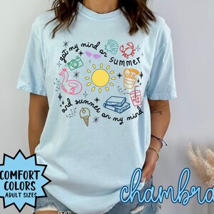 May include: A light blue t-shirt with a graphic design of summer-themed items, including a sun, a flamingo float, a camera, sunglasses, a crab, a seashell, a drink, books, and ice cream. The text on the shirt reads "got my mind on summer and summer on my mind".
