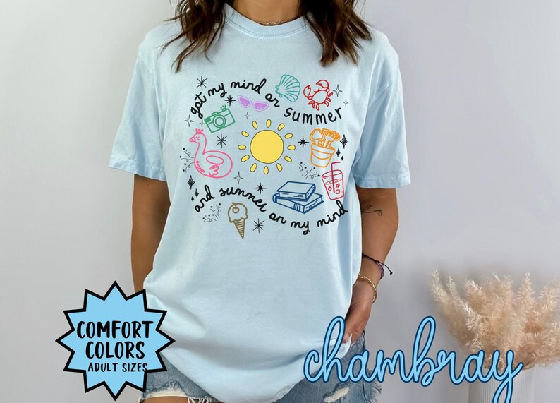 May include: A light blue t-shirt with a graphic design of summer-themed items, including a sun, a flamingo float, a camera, sunglasses, a crab, a seashell, a drink, books, and ice cream. The text on the shirt reads "got my mind on summer and summer on my mind".