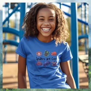 Pode incluir: Uma camiseta azul com um gr&aacute;fico de aranhas de desenho animado e o texto "Spiders are Cool". A camiseta est&aacute; sendo usada por uma pessoa sorrindo, com uma estrutura de playground azul ao fundo.