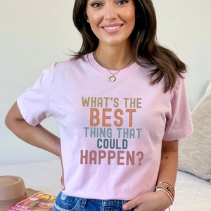 Puede incluir: Una camiseta rosa con el texto "What's the best thing that could happen?" en letras de colores.