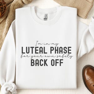 May include: White sweatshirt with the text "I'm in my luteal phase for your own safety back off" printed in black.