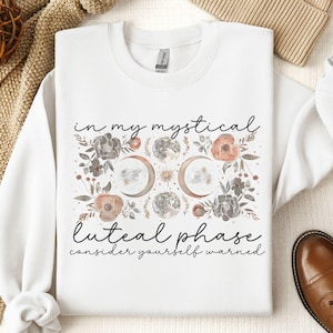 May include: White sweatshirt with a floral design and the text "in my mystical luteal phase consider yourself warned".