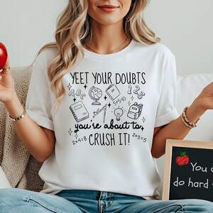 Puede incluir: Camiseta blanca con el texto "YEET YOUR DOUBTS" y gr&aacute;ficos escolares. La camiseta tambi&eacute;n tiene el texto "you're about to CRUSH IT!". Una peque&ntilde;a pizarra muestra el texto "You can do hard things."