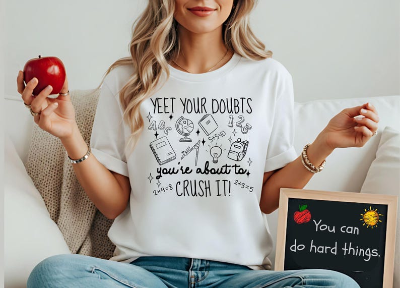 Puede incluir: Camiseta blanca con el texto "YEET YOUR DOUBTS" y gr&aacute;ficos escolares. La camiseta tambi&eacute;n tiene el texto "you're about to CRUSH IT!". Una peque&ntilde;a pizarra muestra el texto "You can do hard things."