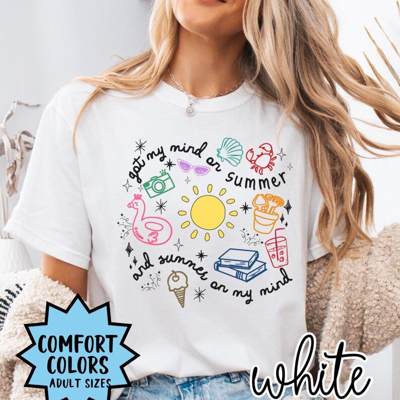 May include: White t-shirt with a colorful graphic design featuring a sun, sunglasses, a beach umbrella, a crab, a camera, a flamingo float, and the text "got my mind on summer and summer on my mind".