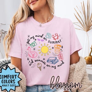 May include: Pink t-shirt with a graphic design of a sun, ice cream, books, and other summer-themed items. The text on the shirt reads "got my mind on summer and summer on my mind".