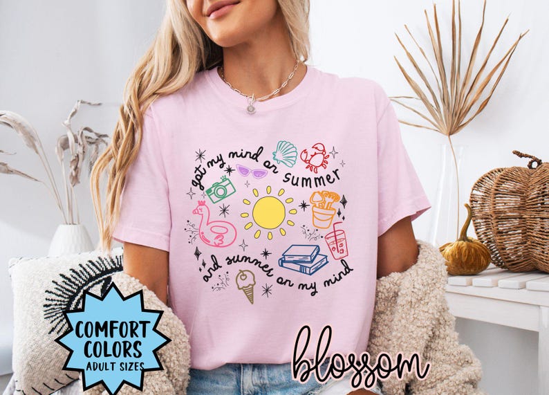 May include: Pink t-shirt with a graphic design of a sun, ice cream, books, and other summer-themed items. The text on the shirt reads "got my mind on summer and summer on my mind".
