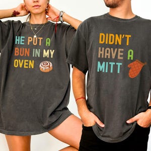 May include: Two charcoal gray t-shirts with matching text and graphics. One shirt reads "HE PUT A BUN IN MY OVEN" with a cinnamon roll graphic. The other shirt says "DIDN'T HAVE A MITT" with an oven mitt graphic.