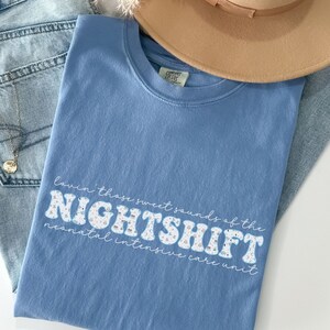 May include: A light blue t-shirt with the text "Lovin' those sweet sounds of the NIGHTSHIFT neonatal intensive care unit" in a retro font. The shirt is paired with light wash jeans and a tan hat.