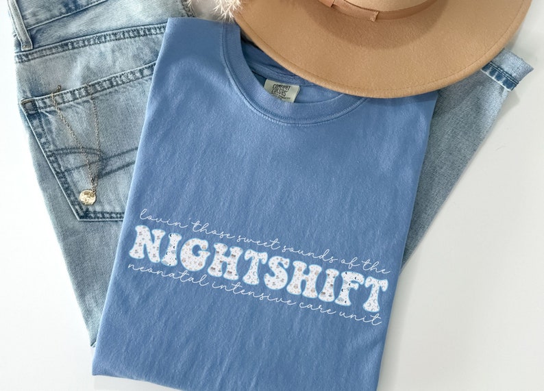 May include: A light blue t-shirt with the text "Lovin' those sweet sounds of the NIGHTSHIFT neonatal intensive care unit" in a retro font. The shirt is paired with light wash jeans and a tan hat.
