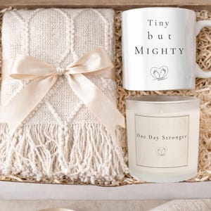 May include: Gift box with a cream-colored blanket tied with a satin ribbon, a white mug with the text "Tiny but Mighty", and a candle with the text "One Day Stronger". The box is filled with straw.