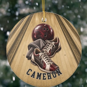 May include: A round ceramic ornament featuring a bowling theme with a dark red bowling ball, a grey towel, and a pair of brown and white bowling shoes resting on a wooden bowling lane. The name CAMERON is printed in dark blue capital letters.