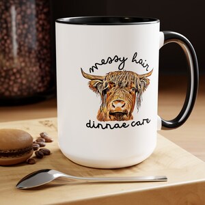 May include: White ceramic mug with a black rim and handle. The mug features a cartoon illustration of a Highland cow with the text "messy hair, don't care".