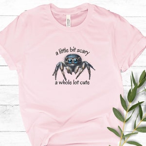 May include: Light pink t-shirt featuring a watercolor illustration of a jumping spider with large blue eyes. The text "a little bit scary, a whole lot cute" is above and below the spider. The shirt is made of soft material.
