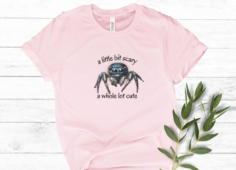 May include: Light pink t-shirt featuring a watercolor illustration of a jumping spider with large blue eyes. The text "a little bit scary, a whole lot cute" is above and below the spider. The shirt is made of soft material.