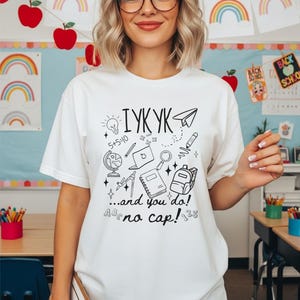 Puede incluir: Camiseta blanca con un gr&aacute;fico negro. El dise&ntilde;o incluye el texto "IYKYK ...and you do! no cap!" y garabatos relacionados con la escuela, como una bombilla, un globo terr&aacute;queo, un port&aacute;til, una mochila y un avi&oacute;n de papel.