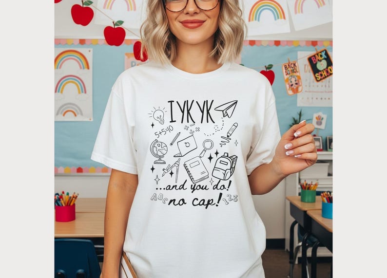 Puede incluir: Camiseta blanca con un gr&aacute;fico negro. El dise&ntilde;o incluye el texto "IYKYK ...and you do! no cap!" y garabatos relacionados con la escuela, como una bombilla, un globo terr&aacute;queo, un port&aacute;til, una mochila y un avi&oacute;n de papel.