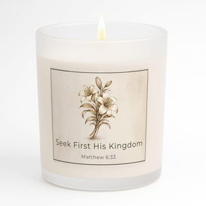 May include: A lit, frosted glass candle with a floral design and the text "Seek First His Kingdom Matthew 6:33." The candle has a beige label with a sepia-toned lily illustration. The flame is bright.