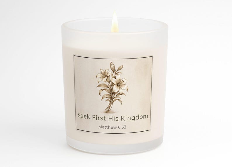 May include: A lit, frosted glass candle with a floral design and the text "Seek First His Kingdom Matthew 6:33." The candle has a beige label with a sepia-toned lily illustration. The flame is bright.