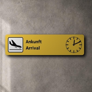 Könnte beinhalten: Ein rechteckiges gelbes Wandschild mit einem schwarzen Flugzeugsymbol auf der linken Seite, dem Text "Ankunft" und "Arrival" in der Mitte sowie einer schwarzen analogen Uhr auf der rechten Seite, deren Zeiger auf etwa zehn nach eins stehen.